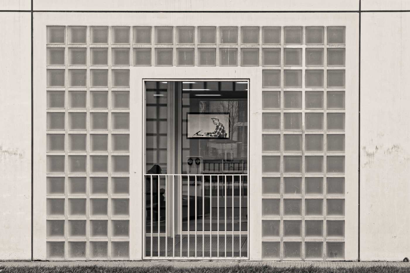 Blick durch das Fenster der Stadtbibliothek Stuttgart auf einen Bildschirm, auf dem eine Präsentation läuft. Erkennbar ist die typische Fassade der Stadtbibliothek aus Glasbausteinen